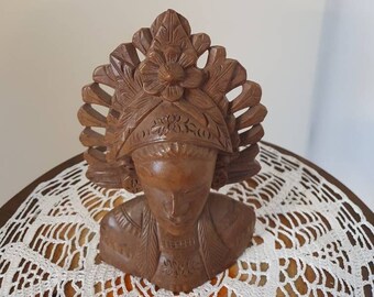 May include: A wooden bust of a woman with a carved headdress. The bust is brown and has intricate details. The woman is wearing a traditional garment.