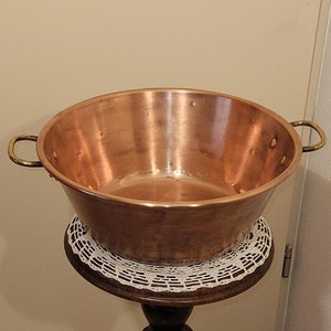 Puede incluir: Un cuenco de cobre con asas de latón se asienta sobre un pedestal de madera con un encaje blanco de ganchillo. El pedestal está tallado con una figura de una persona.
