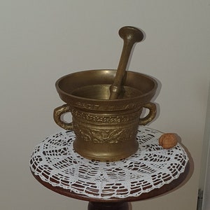 May include: A brass mortar and pestle with intricate designs sits on a white doily atop a wooden pedestal stand with a carved figure.