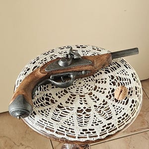 Peut inclure: Un pistolet à silex avec une crosse en bois foncé et des composants métalliques repose sur un napperon en dentelle blanche posé sur un piédestal en bois sculpté. Une noix brune est placée à côté du pistolet sur la dentelle.