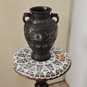 Peut inclure: Un vase noir et or avec des motifs floraux complexes et des accents d'émail colorés repose sur un napperon en crochet blanc sur une table en bois avec une figure sculptée.