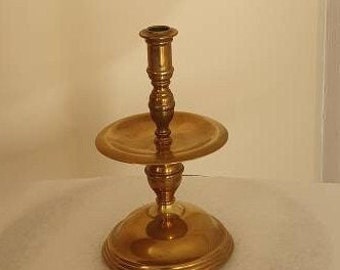 May include: A gold metal candlestick with a round base and a single candle holder. The candlestick has a decorative design on the base and the candle holder.