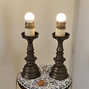 Puede incluir: Dos lámparas de mesa de bronce antiguo con bombillas blancas sobre una mesa de madera con un mantel de ganchillo blanco.