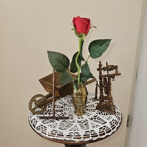 Op de afbeelding: Een decoratieve tafel met een rode roos in een messing vaas. De tafel is versierd met een wit kanten kleedje en miniatuur houten beeldjes, waaronder een kruiwagen en een spinnewiel. De tafelvoet heeft een gesneden figuur.