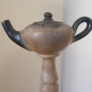 May include: A ceramic teapot with a black glaze and a tall, slender base. The teapot has a round body and a small spout. The base is cylindrical and has a slight bulge in the middle.