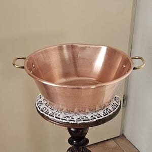 Puede incluir: Un cuenco grande de color cobre con asas de latón. El cuenco es redondo con una abertura ancha y se apoya sobre un soporte de madera decorativo. El soporte tiene un acabado oscuro y un tapete de encaje blanco.