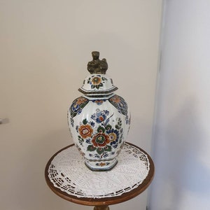 Könnte beinhalten: Eine weiße Keramikvase mit einem Blumendesign in Blautönen, Orange und Grün. Die Vase hat einen goldfarbenen Deckel mit einer kleinen Figurine oben. Die Vase steht auf einer weißen Häkeldeckchen auf einem Holzständer.