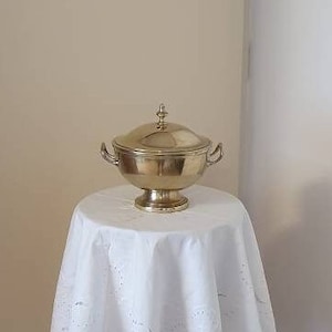 May include: A gold-colored, lidded serving dish with handles, resting on a white tablecloth. The tablecloth is draped over a small, ornate stand. The background is a neutral wall and a white door.