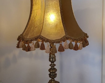 May include: A gold table lamp with a scalloped, fringed lampshade. The lampshade is a combination of gold and brown fabric. The lamp base is ornate and has a decorative design.