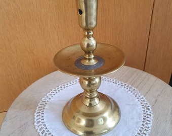May include: A gold metal candlestick with a round base and a tall, slender candle holder. The candlestick is sitting on a white crocheted doily.