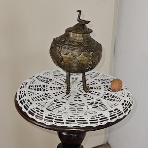 May include: A small, ornate, gold-colored metal pot with a bird-shaped lid sits on a white crocheted doily on a dark wooden table with a carved figure.
