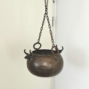 May include: A hanging copper cauldron with a dark interior, suspended by a chain and a black cord. The cauldron has decorative handles and a rounded shape, ideal for decorative purposes.