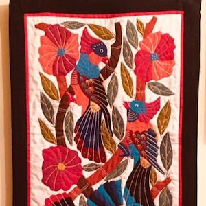 Puede incluir: Una colorida obra de arte textil con pájaros, flores y follaje. La obra presenta pájaros con plumaje azul, rojo y naranja posados en ramas marrones. También hay flores rojas y naranjas y hojas verdes. La pieza está enmarcada con un borde negro.