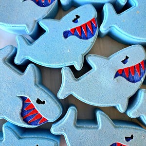 May include: A collection of light blue shark-shaped bath bombs. Each shark features a dark blue mouth with sharp, bright red teeth and a small black eye. The bath bombs are arranged in a cluster, showcasing their uniform shark silhouette.