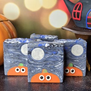 May include: Three rectangular bars of soap featuring a swirling dark blue and black night sky pattern with a white full moon. Each bar displays a bright orange pumpkin base at the bottom with two small white and black circular eyes.