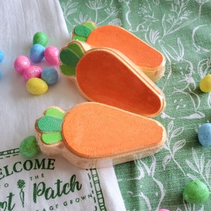 Könnte beinhalten: Drei orangefarbene Badebomben in Form von Karotten mit grünen Spitzen. Die Badebomben liegen auf einem weißen Handtuch mit grünem Muster.