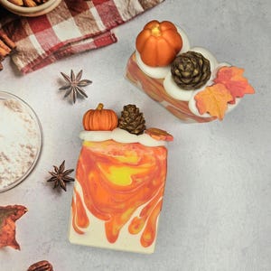 May include: Two rectangular bars of soap featuring a marbled orange, yellow, and white base. Each bar is topped with white soap frosting, a small orange pumpkin, a brown pinecone, and orange and red maple leaf decorations.