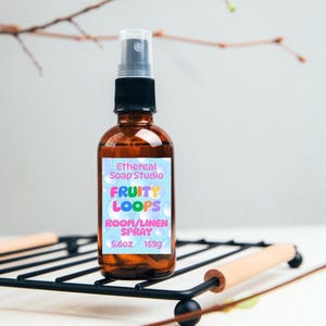 May include: An amber glass spray bottle with a black nozzle, featuring a light blue label with bubble graphics. The label reads Ethereal Soap Studio, FRUITY LOOPS, ROOM/LINEN SPRAY, 5.6oz, and 159g. The bottle is clear and has a cylindrical shape.