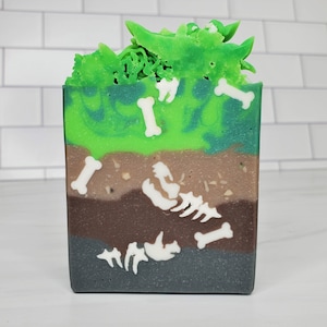 May include: A square bar of handmade soap with layers of color resembling a cross-section of earth. The top layer is bright green with green swirls, followed by brown and gray layers. White bone shapes and a skeleton design are embedded.
