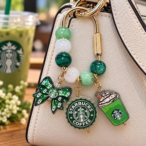 May include: A gold-toned metal keychain featuring a U-shaped clasp adorned with alternating green and white beads. Three charms hang from the clasp: a green bow with small coffee cup icons, a round green Starbucks Coffee logo, and a green iced drink.