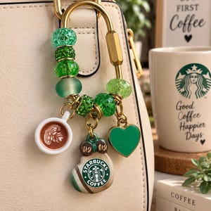 May include: A gold-toned carabiner keychain adorned with various green beads and three charms: a miniature coffee cup with latte art, a heart-shaped green pendant, and a Starbucks coffee-themed charm featuring the brand logo and coffee beans.