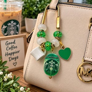 May include: A gold-toned metal carabiner bag charm featuring white and glittery green beads, a miniature white coffee mug charm, a green heart charm, and an oval-shaped green pendant with the Starbucks logo and text printed on the front surface.