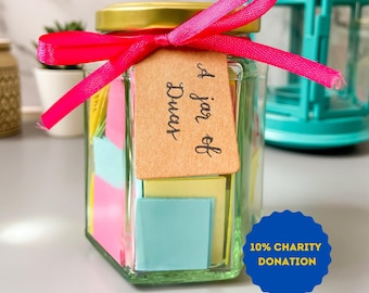May include: A glass jar filled with colourful paper squares tied with a pink ribbon and a brown tag that says "A jar of Dreams". A blue and white lantern is in the background. The sticker on the jar says "10% Charity Donation".