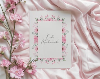 Puede incluir: Una tarjeta blanca con un borde floral rosa y el texto "Eid Mubarak" en caligrafía negra. La tarjeta está sobre un fondo de tela rosa con flores rosas.