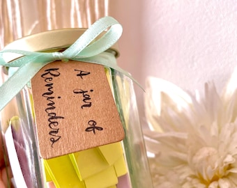 May include: A glass jar filled with colourful paper notes, tied with a light green ribbon and a brown tag that reads "A jar of Reminders".