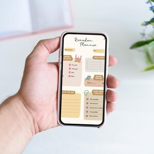 May include: A pink smartphone displaying a digital Ramadan planner with sections for daily goals, prayer checklist, Quran tracker, and good deeds checklist. The planner features a mosque illustration and a light brown background.
