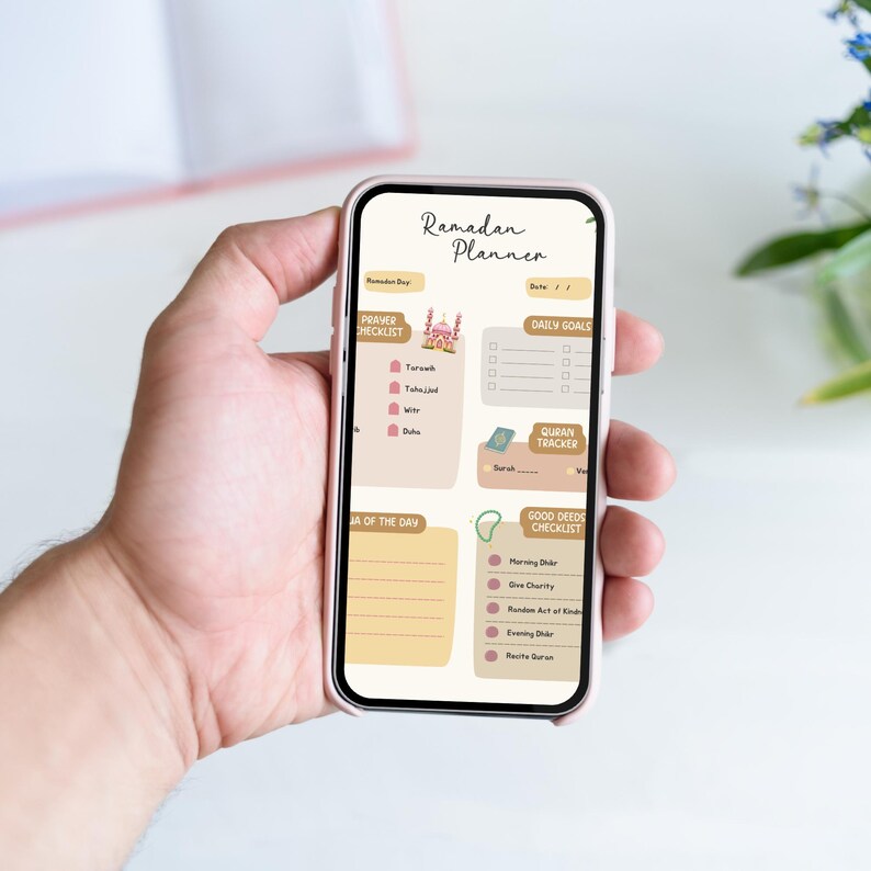 May include: A pink smartphone displaying a digital Ramadan planner with sections for daily goals, prayer checklist, Quran tracker, and good deeds checklist. The planner features a mosque illustration and a light brown background.