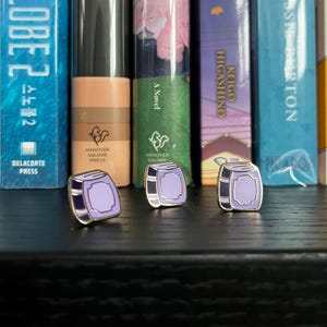 May include: Three enamel pins shaped like books, featuring a purple cover and black spine, with a gold border. They are displayed in front of a bookshelf. The pins are from Hanover Square Press.