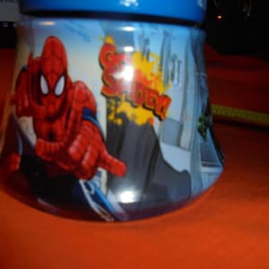 May include: A blue and white ceramic jar featuring the superhero Spider-Man. The jar has a blue lid and a curved shape. The design includes the words "Spider-Man" in red and yellow, and a cityscape background.