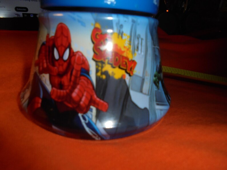 May include: A blue and white ceramic jar featuring the superhero Spider-Man. The jar has a blue lid and a curved shape. The design includes the words "Spider-Man" in red and yellow, and a cityscape background.
