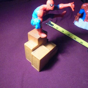 K&ouml;nnte beinhalten: Eine rot-blaue Spider-Man-Actionfigur in hockender Pose auf einem Stapel aus drei braunen Kunststoffkisten. Ein gelbes Ma&szlig;band liegt neben den Kisten und zeigt eine L&auml;nge von etwa 18 cm f&uuml;r die Basisstruktur an.