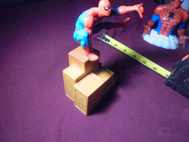 K&ouml;nnte beinhalten: Eine rot-blaue Spider-Man-Actionfigur in hockender Pose auf einem Stapel aus drei braunen Kunststoffkisten. Ein gelbes Ma&szlig;band liegt neben den Kisten und zeigt eine L&auml;nge von etwa 18 cm f&uuml;r die Basisstruktur an.