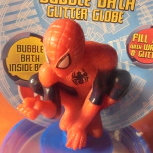 May include: A red and black Spider-Man figurine atop a blue base, part of a bubble bath glitter globe. The packaging features text that reads "Bubble Bath Glitter Globe" and "Fill me with water & glitter."
