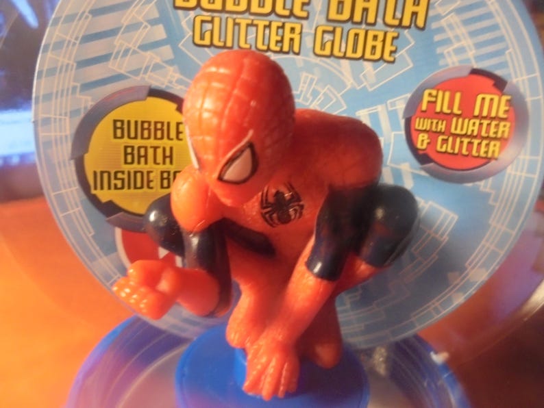 May include: A red and black Spider-Man figurine atop a blue base, part of a bubble bath glitter globe. The packaging features text that reads "Bubble Bath Glitter Globe" and "Fill me with water & glitter."