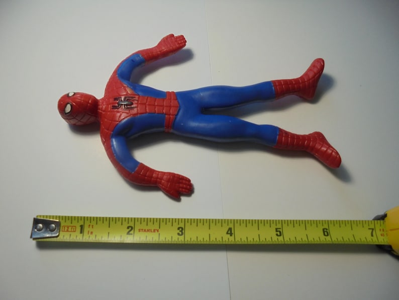 May include: A red and blue Spider-Man action figure with a black spider emblem on the chest. The figure is positioned above a yellow measuring tape, which indicates that the toy measures approximately 17.8 cm in total length from head to toe.