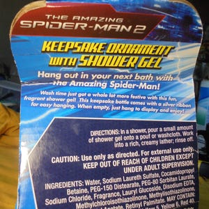 May include: A cardboard product packaging for The Amazing Spider-Man 2 keepsake ornament with shower gel. The blue and red box features white text detailing directions, ingredients, and caution warnings for the fragrant shower gel product.