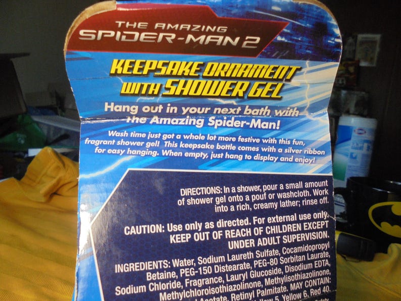 May include: A cardboard product packaging for The Amazing Spider-Man 2 keepsake ornament with shower gel. The blue and red box features white text detailing directions, ingredients, and caution warnings for the fragrant shower gel product.