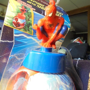 May include: A Spider-Man themed holiday ornament in its original packaging. The ornament features a red and blue Spider-Man figure perched atop a blue cap, which sits on a spherical base decorated with blue and white lightning patterns.