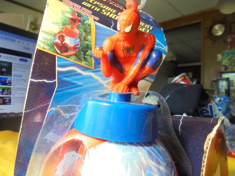 May include: A Spider-Man themed holiday ornament in its original packaging. The ornament features a red and blue Spider-Man figure perched atop a blue cap, which sits on a spherical base decorated with blue and white lightning patterns.
