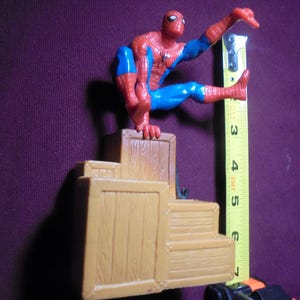 K&ouml;nnte beinhalten: Eine rot-blaue Spider-Man-Actionfigur, die auf einem Stapel beiger Holzkisten sitzt. Ein gelbes Ma&szlig;band ist vertikal neben der Figur positioniert und zeigt eine Gesamth&ouml;he von etwa 17,8 cm f&uuml;r die gesamte Struktur an.