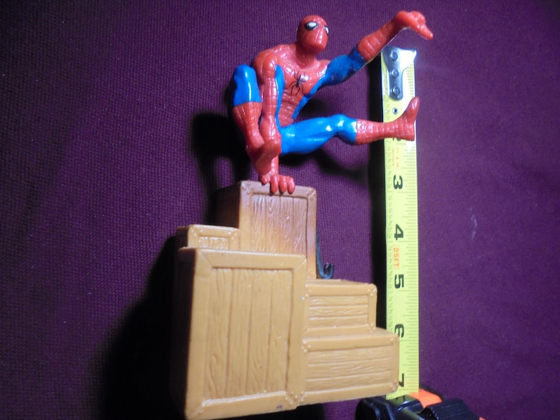 K&ouml;nnte beinhalten: Eine rot-blaue Spider-Man-Actionfigur, die auf einem Stapel beiger Holzkisten sitzt. Ein gelbes Ma&szlig;band ist vertikal neben der Figur positioniert und zeigt eine Gesamth&ouml;he von etwa 17,8 cm f&uuml;r die gesamte Struktur an.
