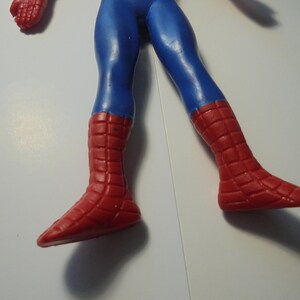 May include: A plastic action figure of a character with blue legs and red boots featuring a raised grid pattern. The figure is shown from the waist down, displaying the blue trousers and the textured red footwear that covers the lower legs and feet.