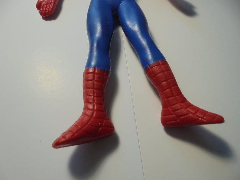 May include: A plastic action figure of a character with blue legs and red boots featuring a raised grid pattern. The figure is shown from the waist down, displaying the blue trousers and the textured red footwear that covers the lower legs and feet.