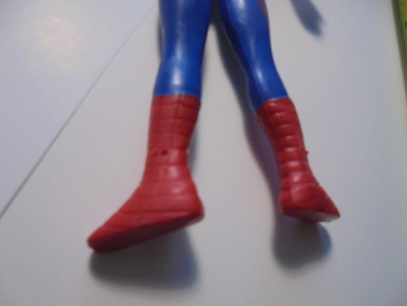 May include: A plastic action figure showing the lower body of a character with blue legs and red boots. The boots feature a textured grid pattern across the surface. The figure is positioned against a plain white background with a partial view of a ruler.