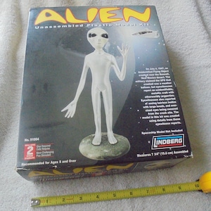 May include: A Lindberg Alien unassembled plastic model kit box featuring a white extraterrestrial figure on a base. The box displays the text Alien, No. 91004, and historical Roswell information. A yellow measuring tape shows the model is 7.75 inches.