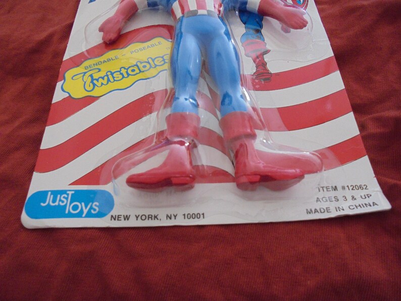 May include: A Captain America action figure in a plastic package. The figure is wearing a blue and red costume with red boots and gloves. The packaging has red and white stripes and the words "Bendable Poseable Twistables".